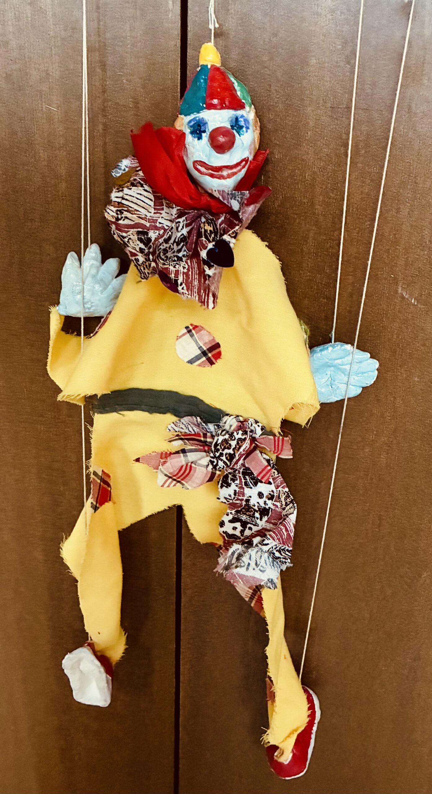 色柄の布の衣装を着たマリオネットが頭と手足に紙粘土のパーツを付けられ、上部の角材から伸びた凧糸で釣られて立っている全身の様子が写っている縦長の写真
